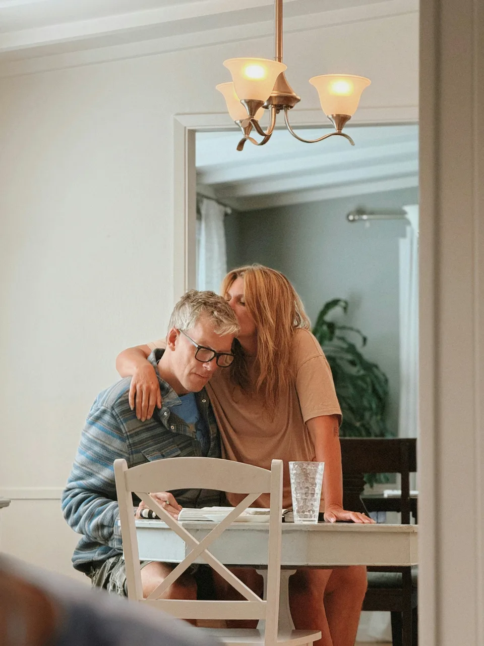 A couple at their kitchen table. She kisses the top of his head while he looks over their notes.